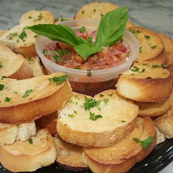 A platter of toasted Italian bread slices with a container of fresh bruschetta topped with basil.