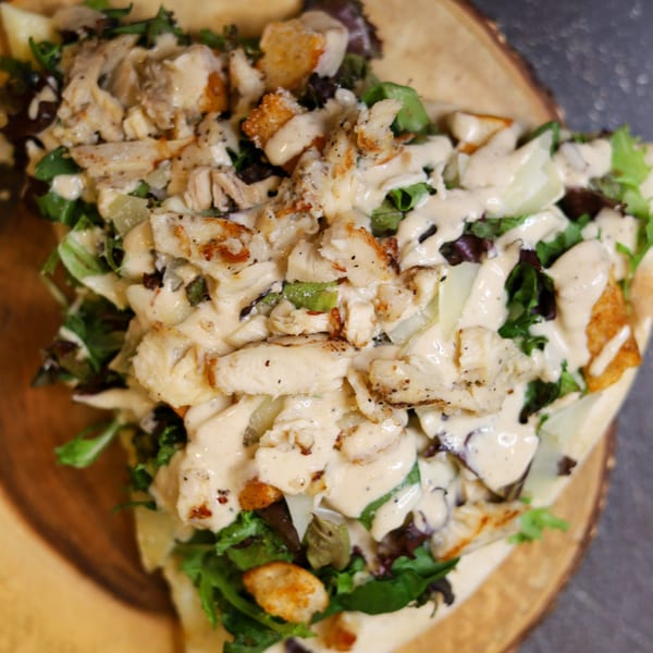 A close-up of a chicken Caesar salad with dressing, croutons, and greens on a wooden board.