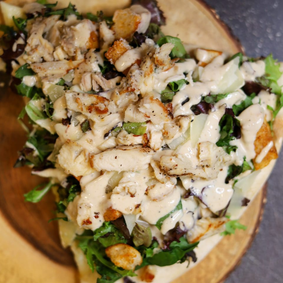 A close-up of a chicken Caesar salad with dressing, croutons, and greens on a wooden board.